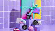 A promotional image for the 'And Breathe' Lush gift set, showcasing all ten included products. The ombré gift box is displayed on a raised purple block, while the products are elegantly arranged on smaller matching blocks below. The backdrop features glossy purple-and-white bathroom tiles, creating a fresh, spa-like atmosphere