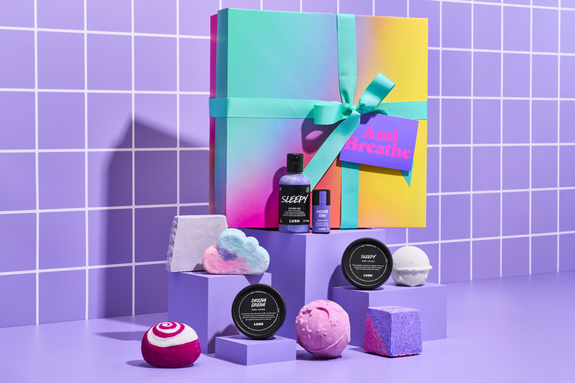 A promotional image for the 'And Breathe' Lush gift set, showcasing all ten included products. The ombré gift box is displayed on a raised purple block, while the products are elegantly arranged on smaller matching blocks below. The backdrop features glossy purple-and-white bathroom tiles, creating a fresh, spa-like atmosphere