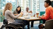 Four people, including a woman in a wheelchair, sit around a table having a discussion in a modern office with large windows and city buildings visible outside.