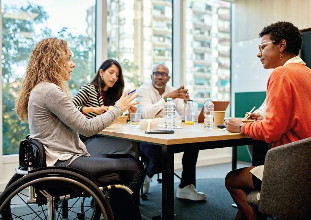 Four people, including a woman in a wheelchair, sit around a table having a discussion in a modern office with large windows and city buildings visible outside.