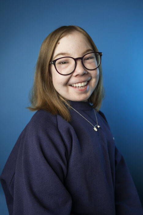 Bella smiles with light brown hair and dark-rimmed glasses wears a navy blue sweater against a vibrant blue background.