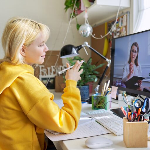 A student watches a teacher on a laptop screen. She holds a pen in her hand ready to take notes.