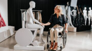 Sophie Morgan, sits in her wheelchair and smiles warmly as she holds the hand of her seated wheelchair mannequin. The mannequin is positioned on a small display platform. Behind them are clothing racks and additional mannequins in various poses. The setting appears to be a showroom or studio space with professional lighting equipment visible in the background.