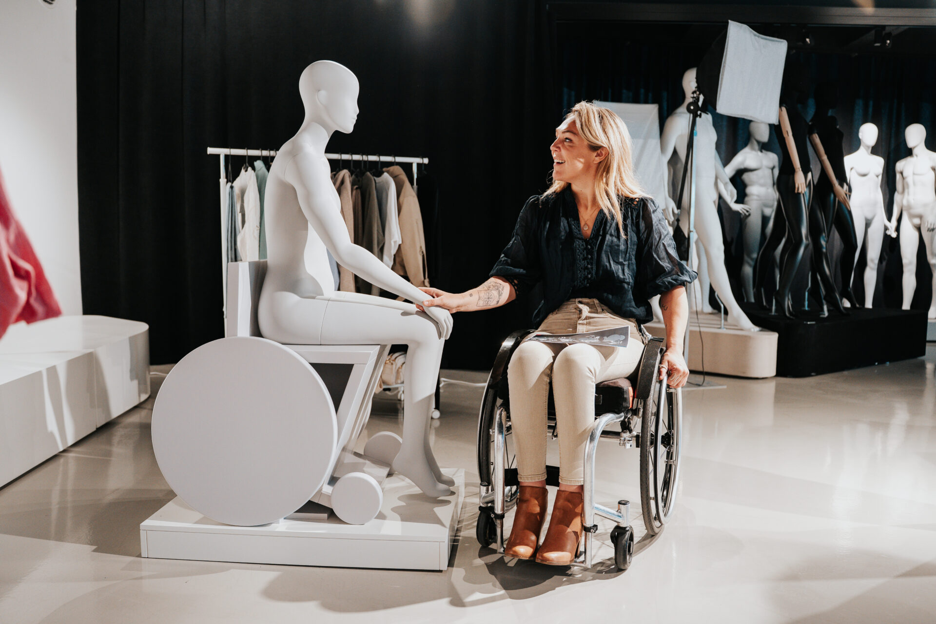 Sophie Morgan, sits in her wheelchair and smiles warmly as she holds the hand of her seated wheelchair mannequin. The mannequin is positioned on a small display platform. Behind them are clothing racks and additional mannequins in various poses. The setting appears to be a showroom or studio space with professional lighting equipment visible in the background.