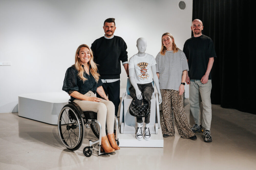 A group of four people pose beside a seated wheelchair mannequin displayed on a small platform. Sophie Morgan sits in her own wheelchair at the left, smiling. The mannequin is dressed in casual clothing, including a graphic T-shirt and sneakers, and holds a studded handbag. Two people stand behind the mannequin, and another person stands to the right. They are pictured in an indoor showroom or exhibition space.