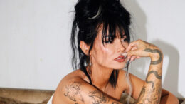 This is a photo of Talia Mar, sitting on a couch, posing to the camera. Her dark hair is styled in a messy updo and she rests her hand near her mouth. She wears hoop earrings, a white strapless top, and has many tattoos covering her arms, including snakes, flowers, and abstract designs. The background is simple and white.