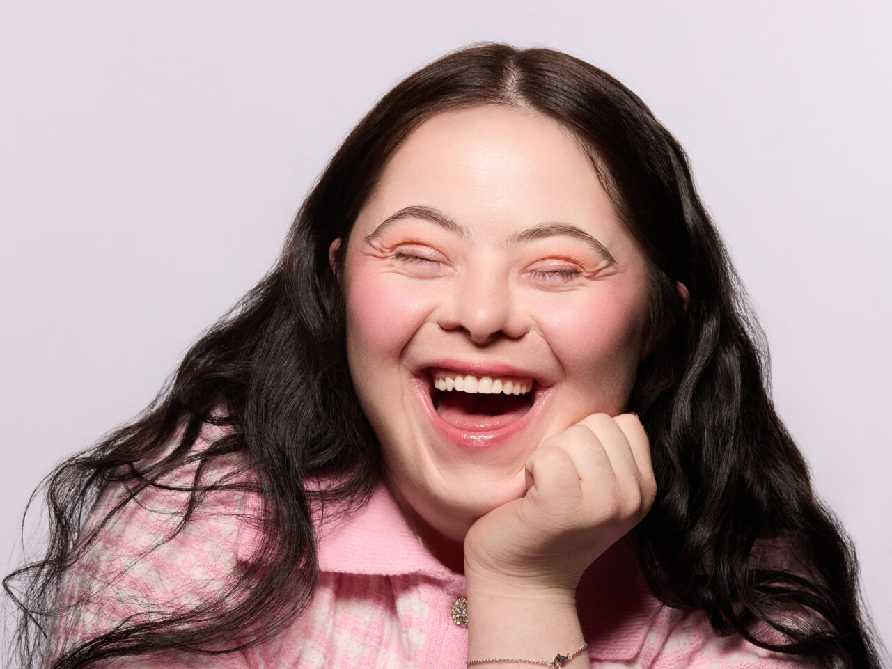 A close up of Ellie Goldstein smiling and laughing with her eyes closed. She is wearing a pink textured top, and rests her hand on her chin.