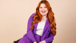 Jenni sits in her chair and smiles at the camera. She is wearing a bright purple suit and her ginger hair lies curled around her shoulders. She appears to be in a studio setting with a soft, beige background.