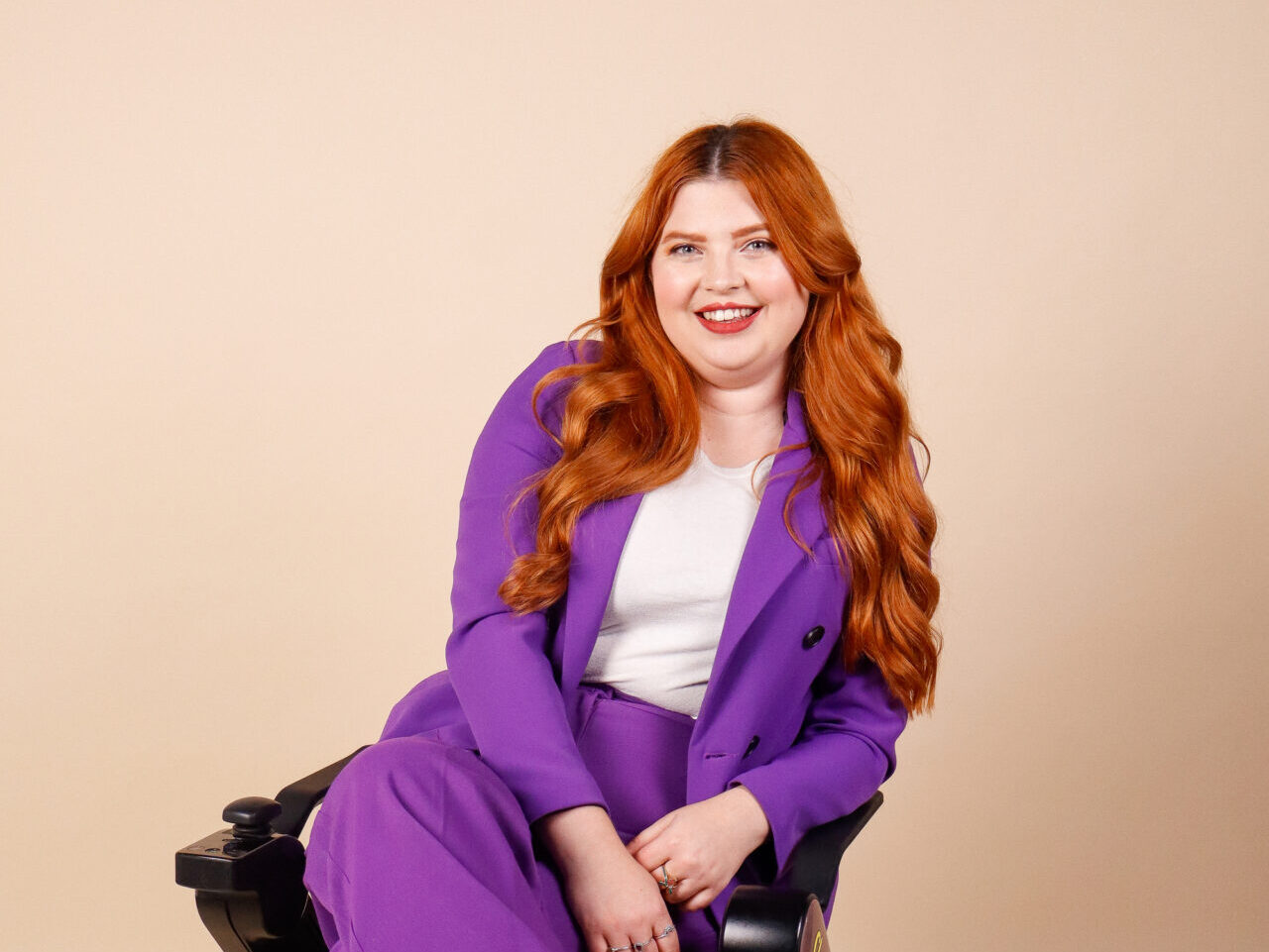 Jenni sits in her chair and smiles at the camera. She is wearing a bright purple suit and her ginger hair lies curled around her shoulders. She appears to be in a studio setting with a soft, beige background.