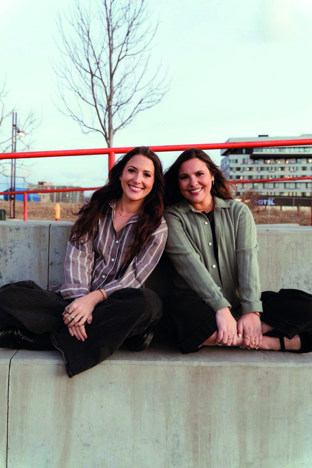 An image of Alexa and Jacqueline Child. They are smiling, both have long brown hair, and are sitting crosslegged against a concrete wall. Alexa is wearing a striped grey shirt and black jeans. Jacqueline is wearing an olive green cardigan and black trousers. They are both wearing rings and bracelets.