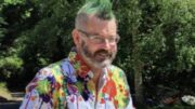 An image of Mark Webb outside on a patio near trees on a sunny day. He is smiling, wearing glasses and has a spiky green mohawk and a dark grey beard. He is wearing a blue shirt with colourful paint blotched over the shoulders, collar, and around the waist. He is using a walking frame.