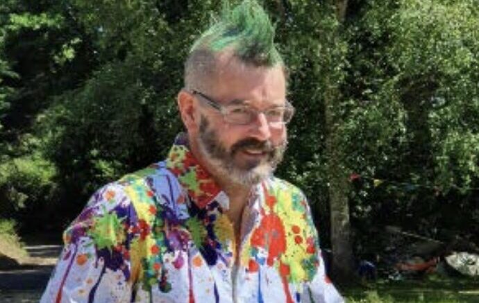 An image of Mark Webb outside on a patio near trees on a sunny day. He is smiling, wearing glasses and has a spiky green mohawk and a dark grey beard. He is wearing a blue shirt with colourful paint blotched over the shoulders, collar, and around the waist. He is using a walking frame.
