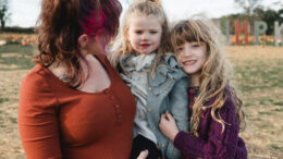 An image of mum Rhiannon and her two daughters Poppy and Charlotte. Rhiannon has brown hair with dyed pink highlights. She is wearing a red top and is holding Poppy in her arms. Poppy is smiling, has blond hair and is wearing a grey cardigan. Charlotte is hugging them and smiling. She has long blonde hair and is wearing a purple knitted jumper.