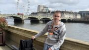 An image of Liam Virgo standing in front of the river Thames with the London Eye and Westminster Bridge in the background. He is smiling, has a buzz cut, and is wearing a grey London hoodie and blue Adidas joggers. He is standing next to a wheel chair and has an orange visitor pass around his neck.