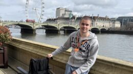 An image of Liam Virgo standing in front of the river Thames with the London Eye and Westminster Bridge in the background. He is smiling, has a buzz cut, and is wearing a grey London hoodie and blue Adidas joggers. He is standing next to a wheel chair and has an orange visitor pass around his neck.