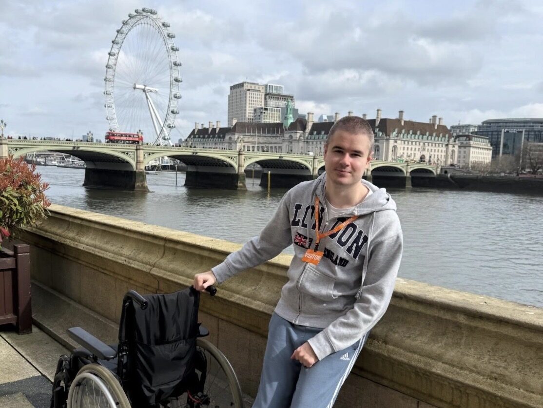 An image of Liam Virgo standing in front of the river Thames with the London Eye and Westminster Bridge in the background. He is smiling, has a buzz cut, and is wearing a grey London hoodie and blue Adidas joggers. He is standing next to a wheel chair and has an orange visitor pass around his neck.