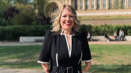 An Image of Dr Marie Tidball MP standing outside the Houses of Parliament. She is smiling and has long blond hair. She is wearing a smart black business dress with white details.