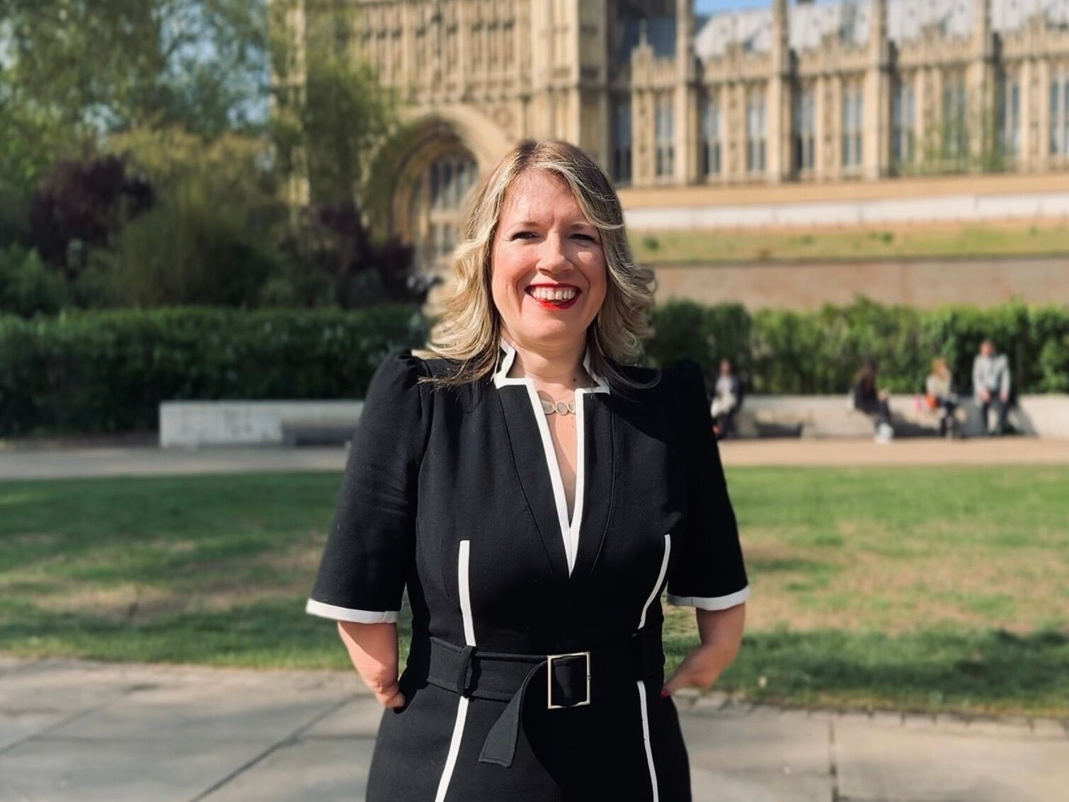 An Image of Dr Marie Tidball MP standing outside the Houses of Parliament. She is smiling and has long blond hair. She is wearing a smart black business dress with white details.