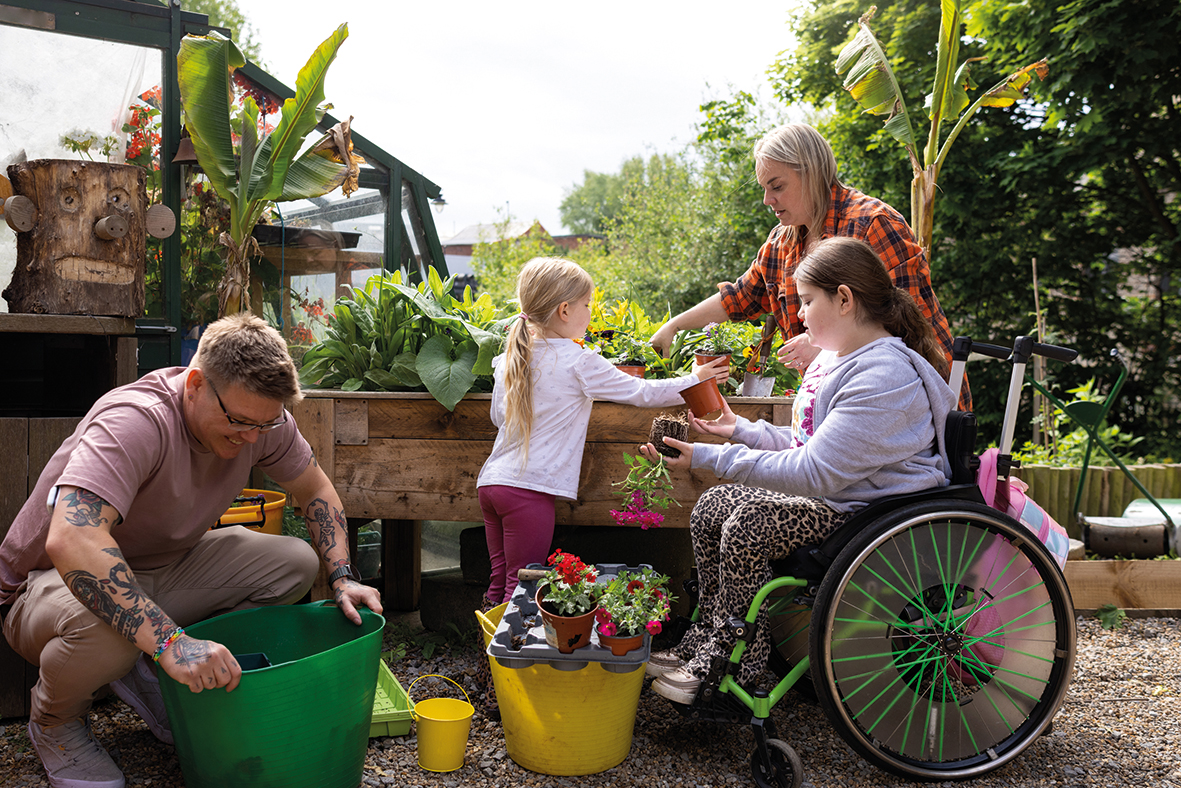 An image of a family spending time in their garden. There are two girls and a mum and dad all working together. One girl is using a wheelchair while repotting a plant.