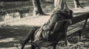 A black and white image of a woman sitting on a park bench. She has long curly blond hair and is wearing a long jacket and scarf. She is facing away from camera.