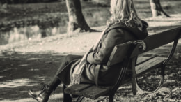 A black and white image of a woman sitting on a park bench. She has long curly blond hair and is wearing a long jacket and scarf. She is facing away from camera.
