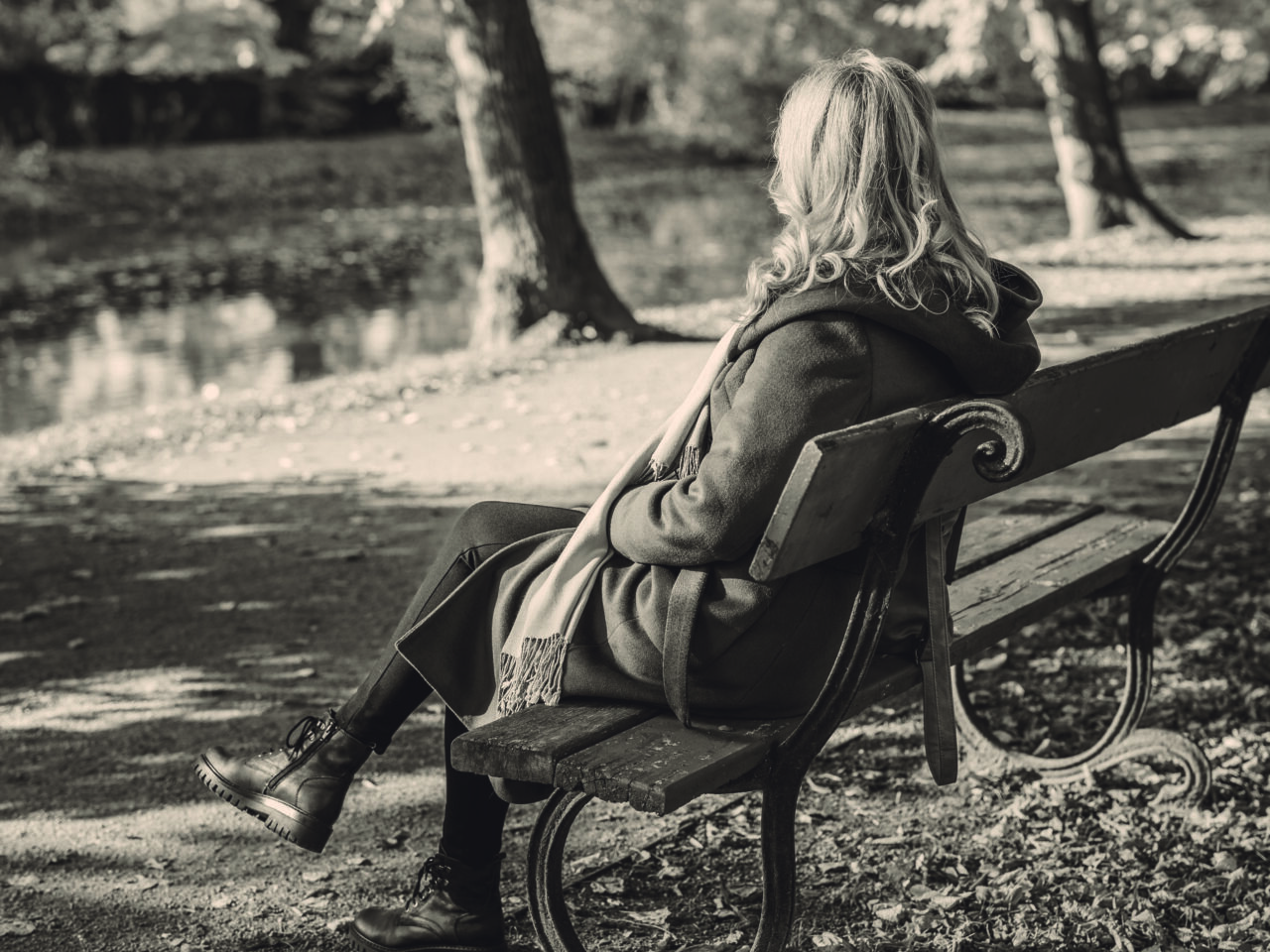 A black and white image of a woman sitting on a park bench. She has long curly blond hair and is wearing a long jacket and scarf. She is facing away from camera.