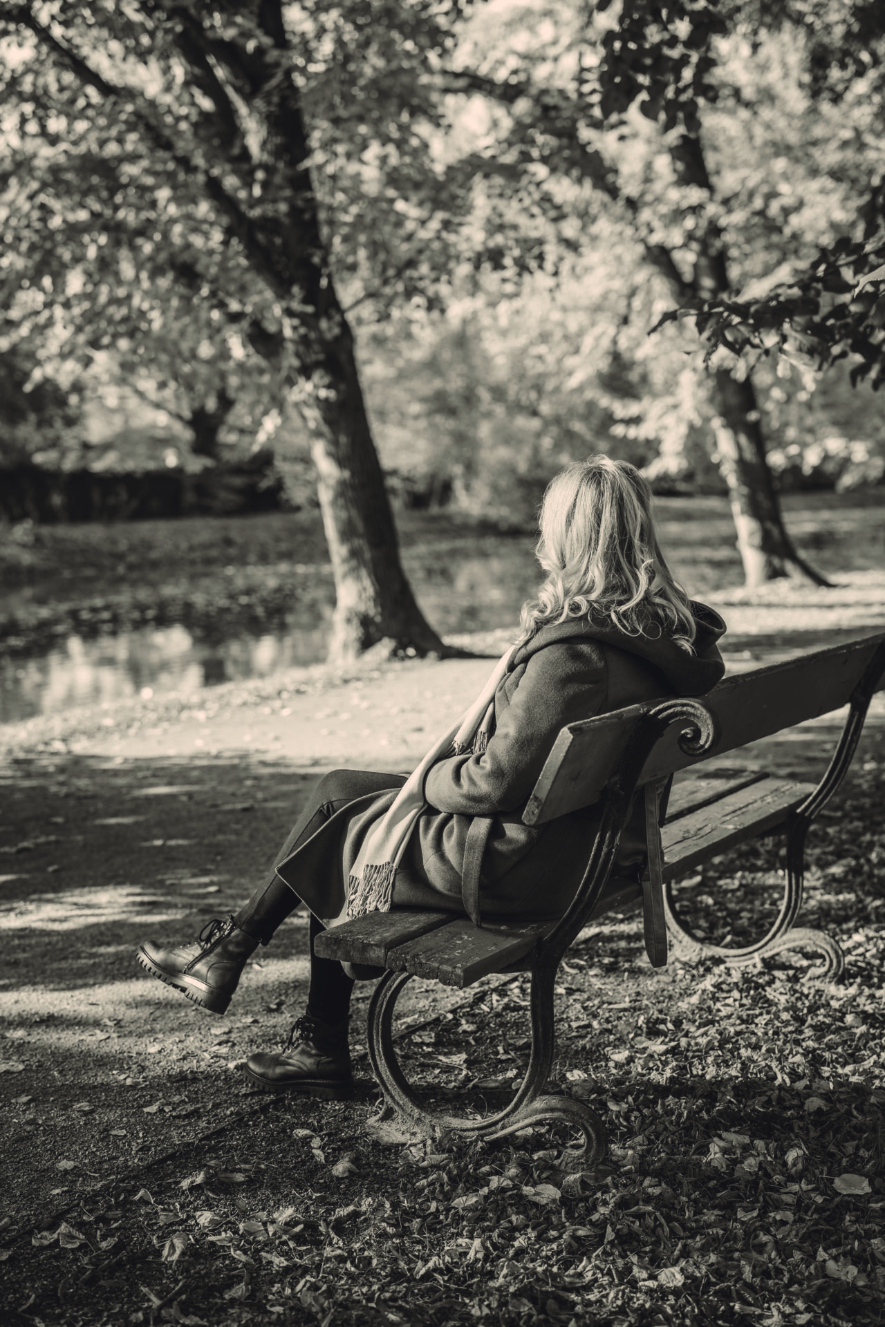 A black and white image of a woman sitting on a park bench. She has long curly blond hair and is wearing a long jacket and scarf. She is facing away from camera.