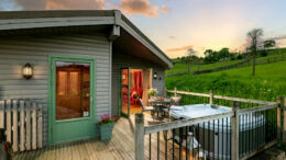A Hoe Grange Holidays accessible cabin pictured at dusk with a large patio in front containing a hot tub and a table and chairs.