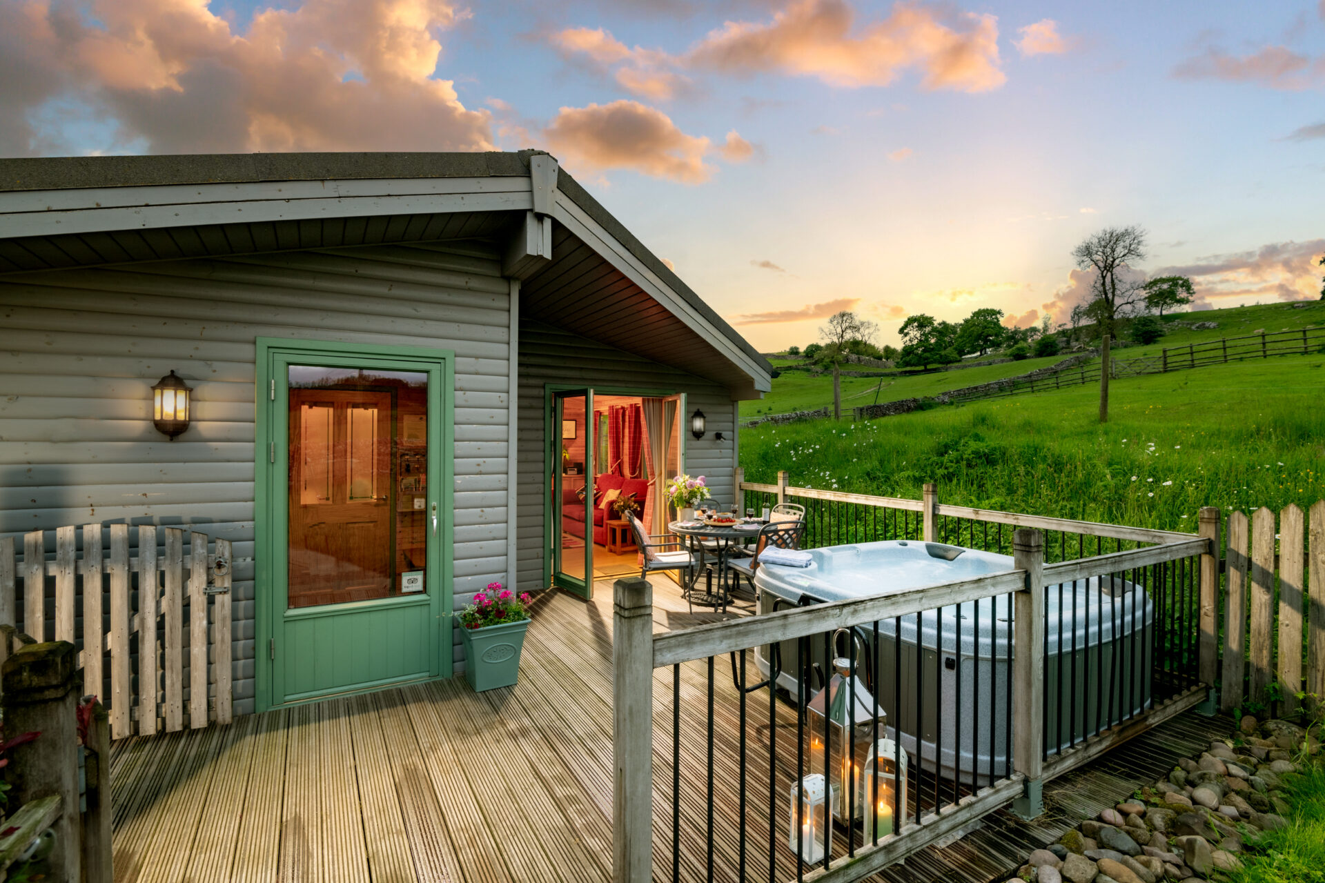 A Hoe Grange Holidays accessible cabin pictured at dusk with a large patio in front containing a hot tub and a table and chairs.