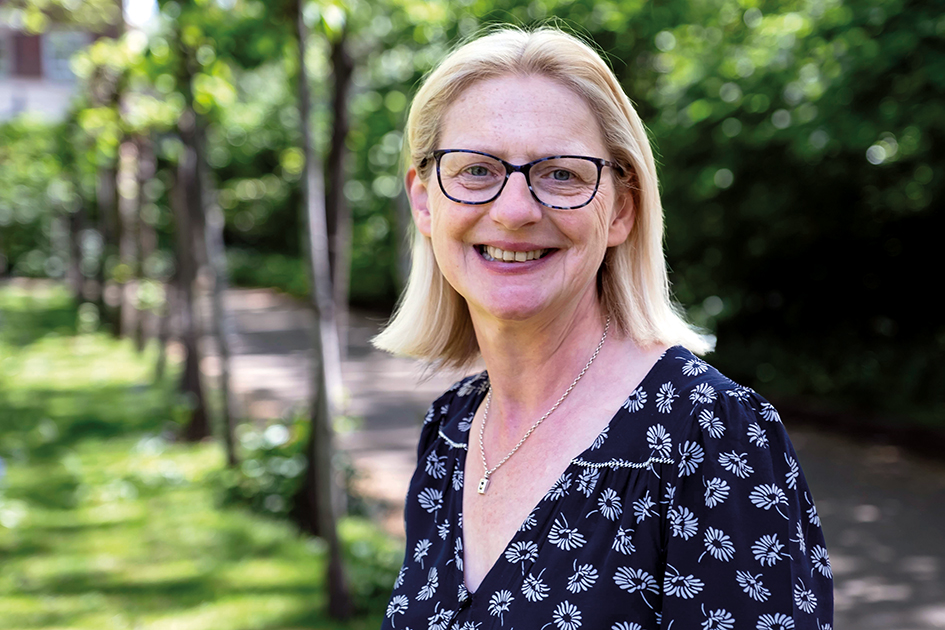 An image of Helen Walker. She is smiling, is wearing glasses and has long blond hair. She is wearing a dark blue floral dress and is pictured outside in a green wooded area on a sunny day.