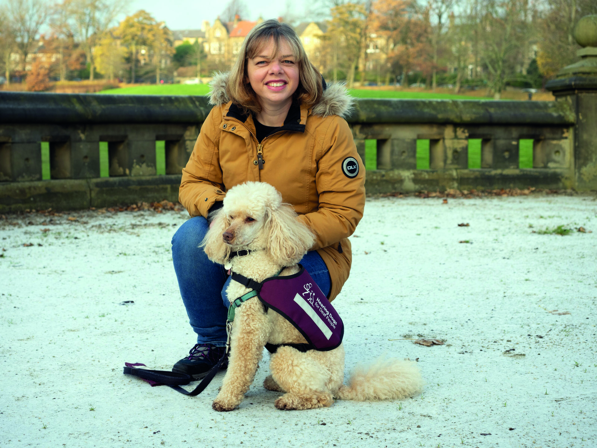 An image of Jenny Jones and her assistance poodle Xander. She is smiling and wearing a yellow winter jacket. She is kneeling behind Xandar who is looking very smart in his burgundy jacket.