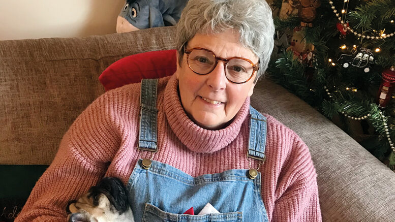 An image of Liz and her pug Jarvis. Liz is smiling, has glasses and grey hair, and is wearing a pink knitted turtleneck and denim overalls. She is holding Jarvis and sitting on a sofa with a Christmas tree in the background.