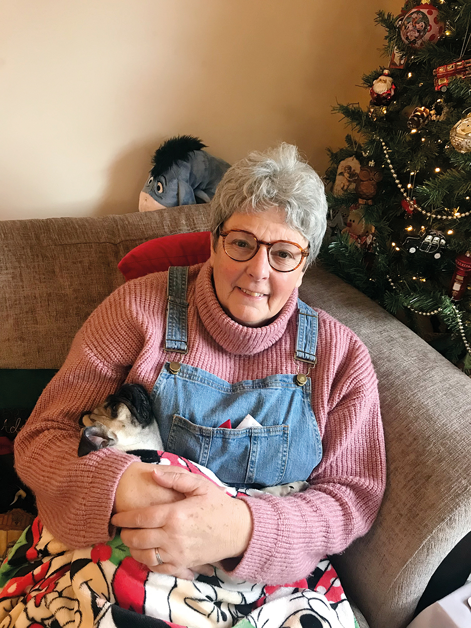 An image of Liz and her pug Jarvis. Liz is smiling, has glasses and grey hair, and is wearing a pink knitted turtleneck and denim overalls. She is holding Jarvis and sitting on a sofa with a Christmas tree in the background.