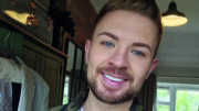 An image of Luke Manton. He is smiling, has short brown hair and a circle beard, and is wearing a dark green and black chequered flannel and a white t-shirt.