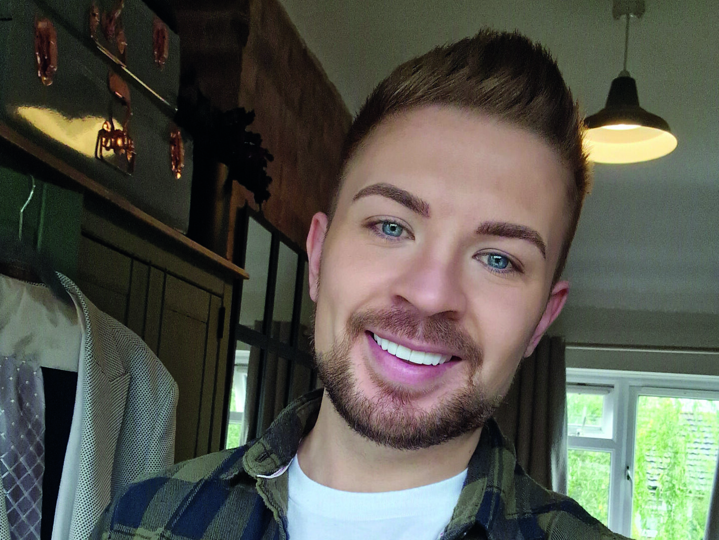An image of Luke Manton. He is smiling, has short brown hair and a circle beard, and is wearing a dark green and black chequered flannel and a white t-shirt.