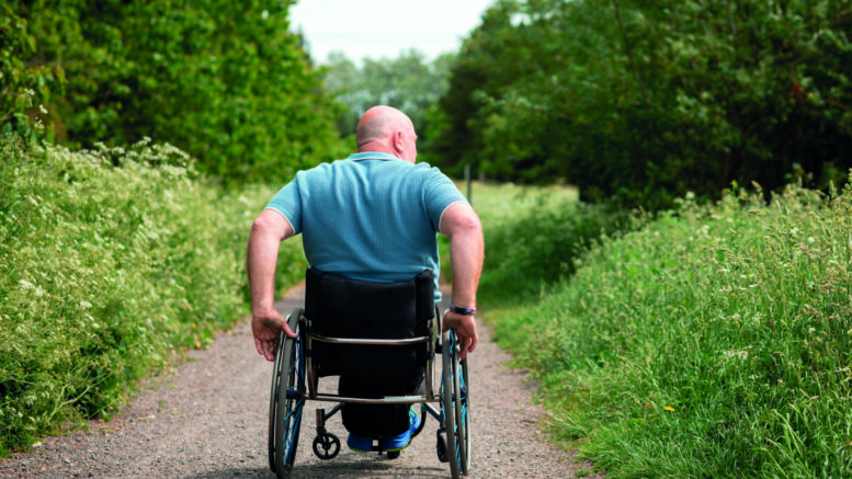 An image of a bald man using a wheelchair on a countryside path surrounded by grass and trees. He is wearing a blue polo shirt and moving away from camera.