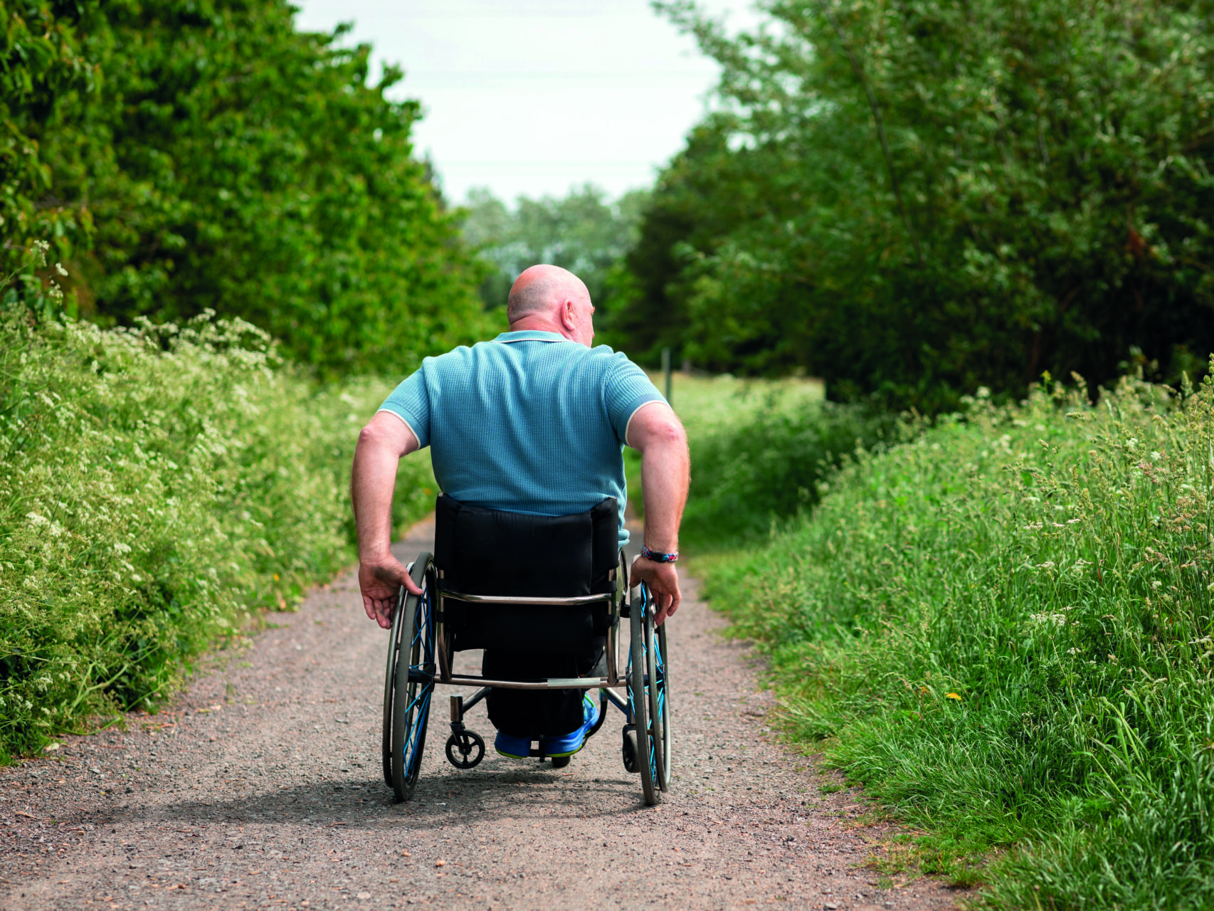 An image of a bald man using a wheelchair on a countryside path surrounded by grass and trees. He is wearing a blue polo shirt and moving away from camera.