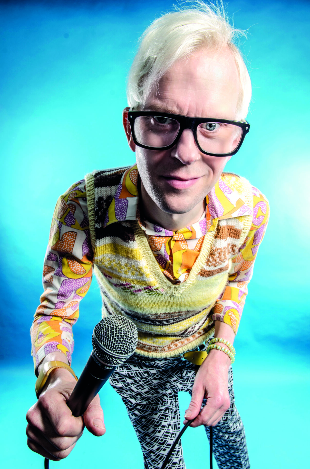 An image of Robert White. He is smiling, has white hair and is wearing glasses. He is holding a microphone and wearing a bright patterned shirt and yellow knitted cardigan.