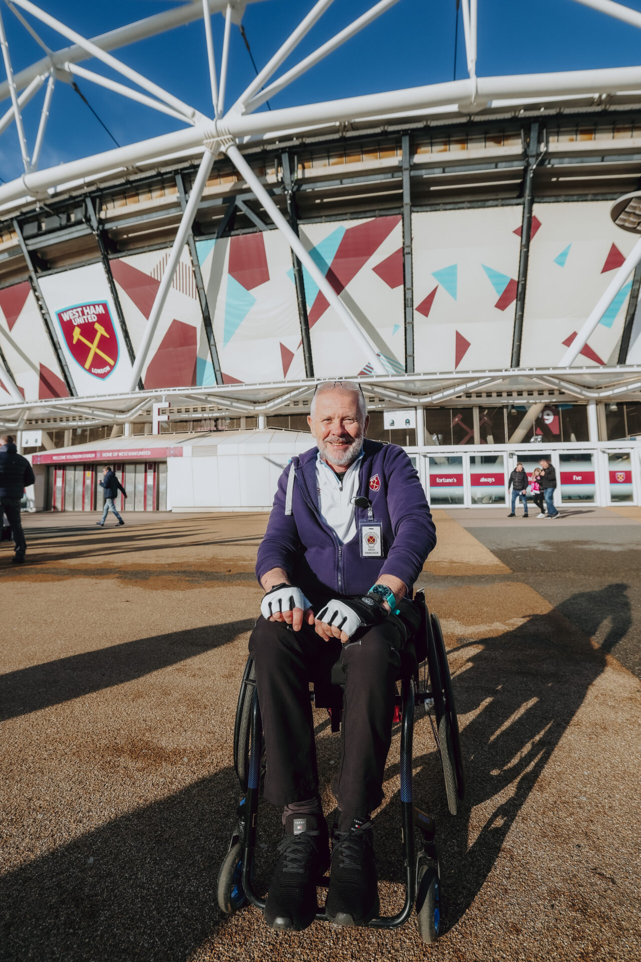 An image of a member of West Ham United's Disabled Supporters' Association. He is smiling, has a white beard and glasses on his forehead, and is using a wheelchair. Behind him is London Stadium.
