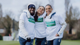An image of three women on a football pitch. They are smiling and wearing white 'unite for access' sports tops and navy blue joggers and shorts.