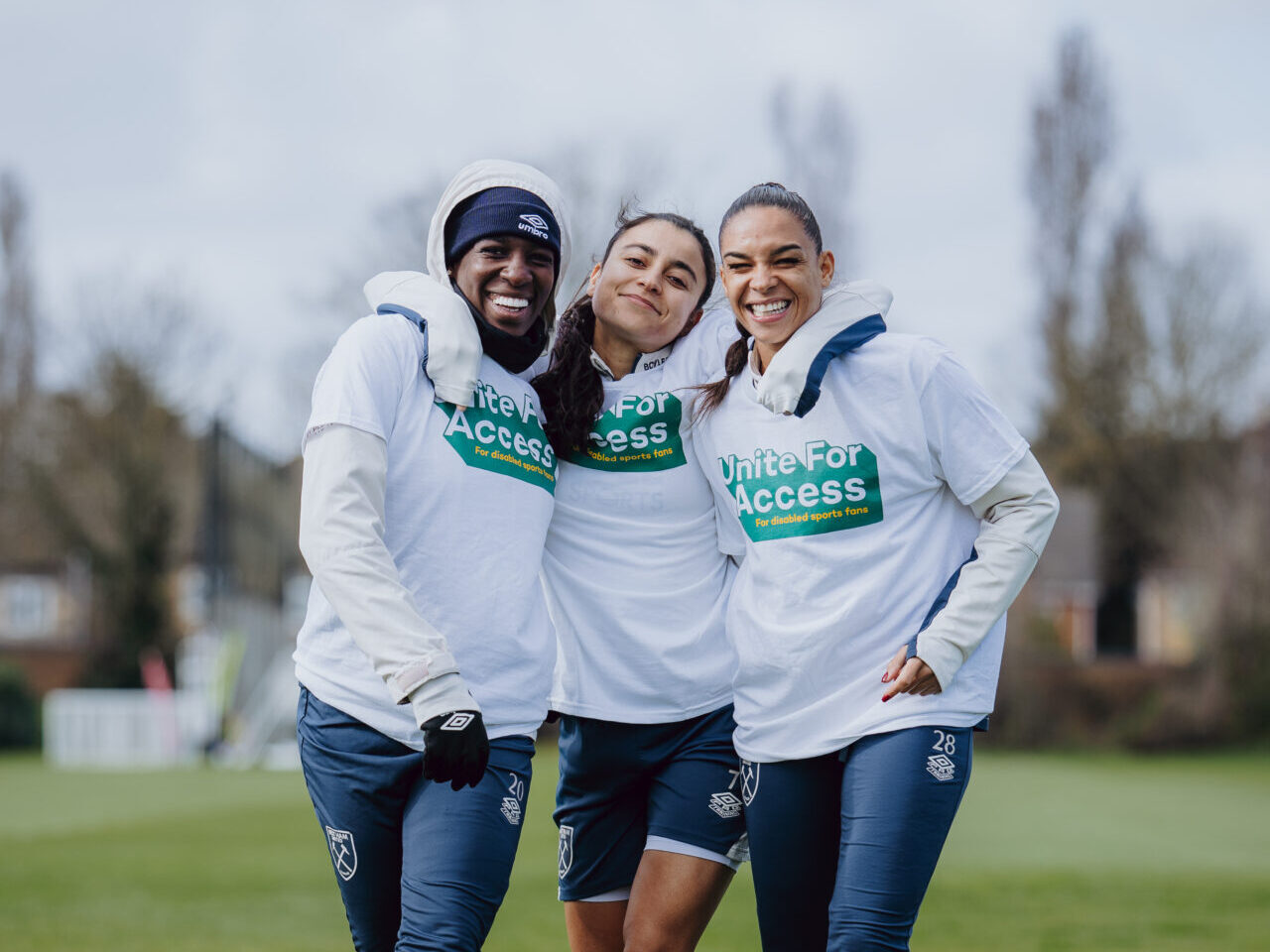 An image of three women on a football pitch. They are smiling and wearing white 'unite for access' sports tops and navy blue joggers and shorts.