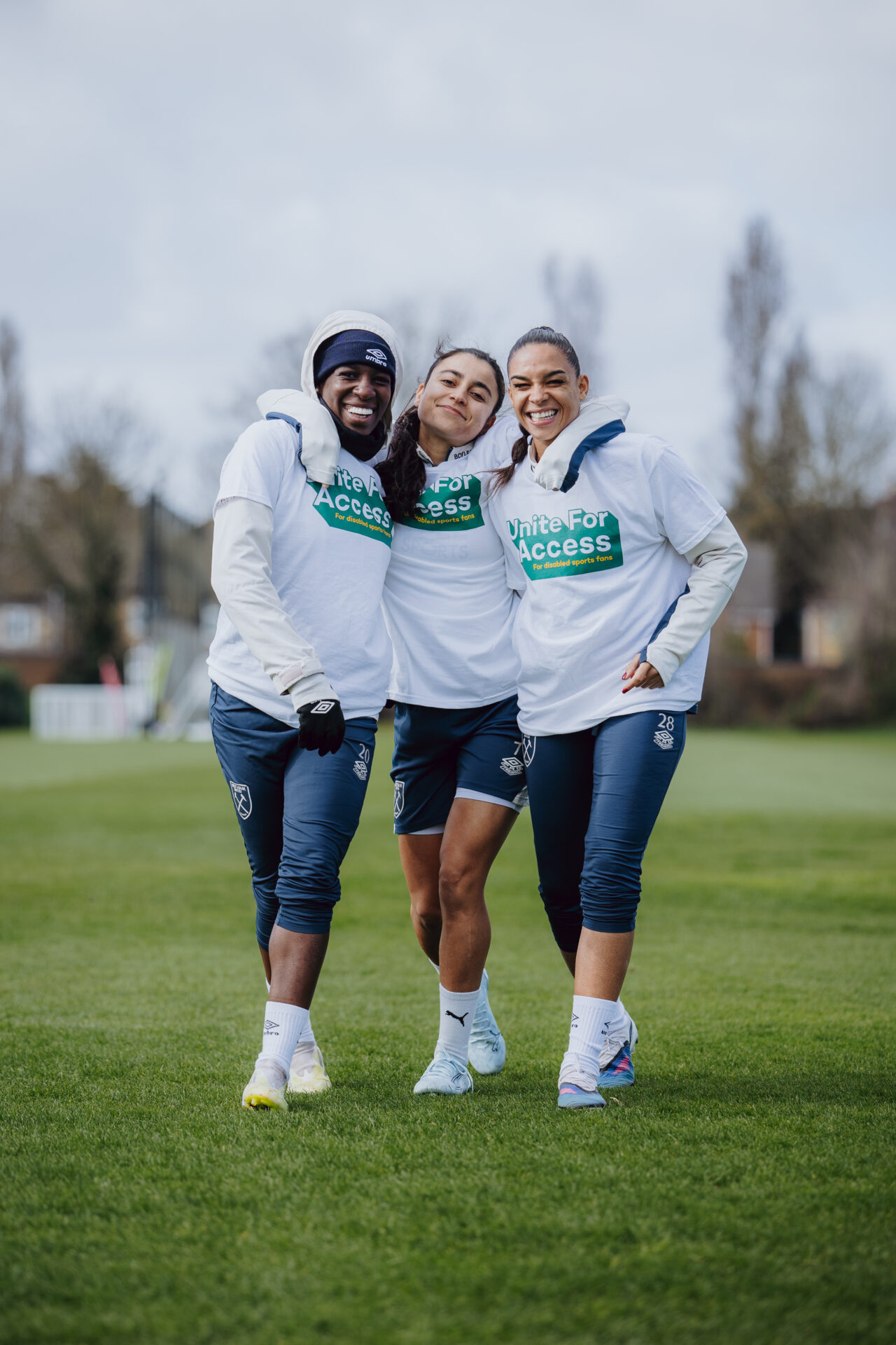 An image of three women on a football pitch. They are smiling and wearing white 'unite for access' sports tops and navy blue joggers and shorts. 