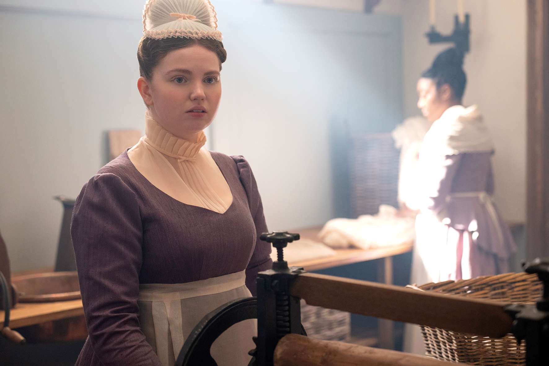 Young woman in historical costume stands in a vintage laundry room with soft lighting, wearing a maid's outfit and cap, while another woman works in the background near baskets and wooden tables.