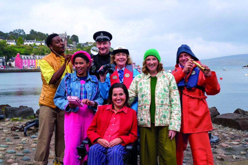 An image of the cast of Balamory. From left to right; Dr Ollie, played by Carl Spencer, who is wearing a gold gilet and yellow top and using a stethoscope; Ava Potts, played by Danielle Jam, who is wearing a bright pink beret, a denim jacket and pink trousers, and holding a remote control; PC Plum played by Andrew Agnew, who is wearing a police officer's uniform and holding a magnifying glass; Edie McCredie played by Juliet Cadzow, who is wearing a blue coat and red life jacket, and using binoculars; Penny Pocket played by Kim Tserkezie who is wearing a red cardigan and shirt, striped blue trousers, and using a wheelchair; Miss Hollie played by Julie Wilson Nimmo who is wearing a green beanie, floral jacket, and a green dress; and the Harbourmaster played by William Andrews, who is wearing a bright orange waterproof jacket, a blue fisherman's hat and life jacket, and is holding a small, gold telescope.