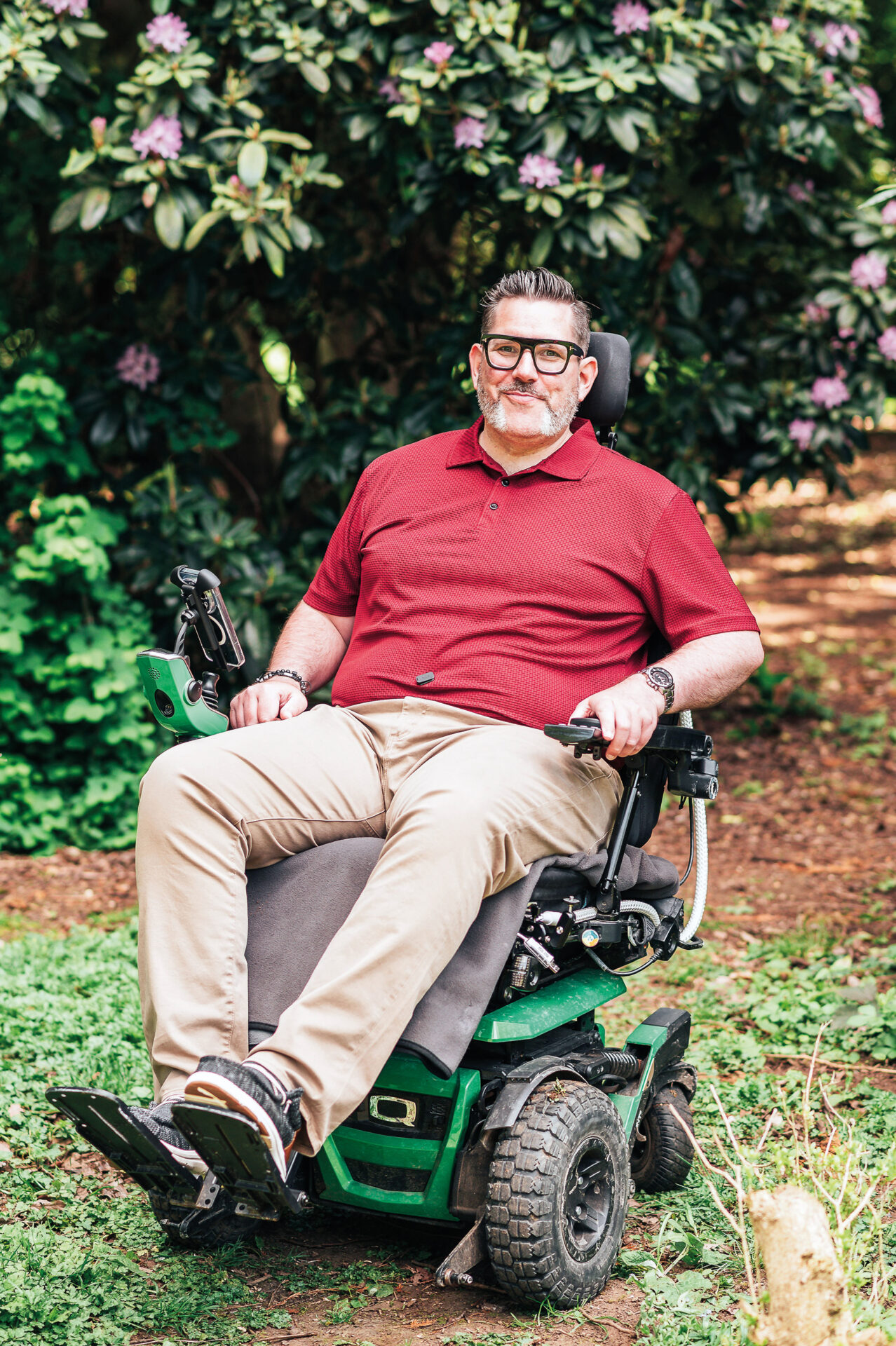 Nick is pictured using a green coloured wheelchair on an outdoor trail. He has short black gelled-up hair and black and grey muttonchops. He is smiling and wearing glasses, a red polo shirt and tan trousers.