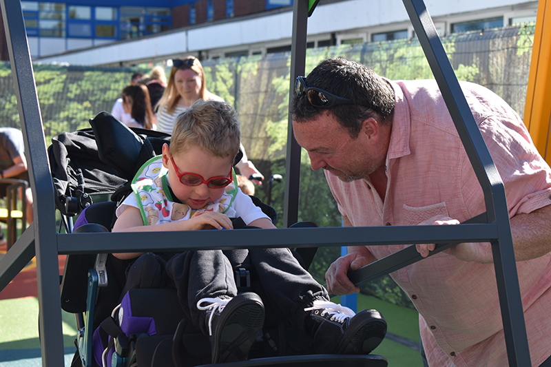 A Fox Wood pupil enjoying using a wheelchair accessible swing, as a man in a pink shirt stands next to him, smiling and pushing the swing.