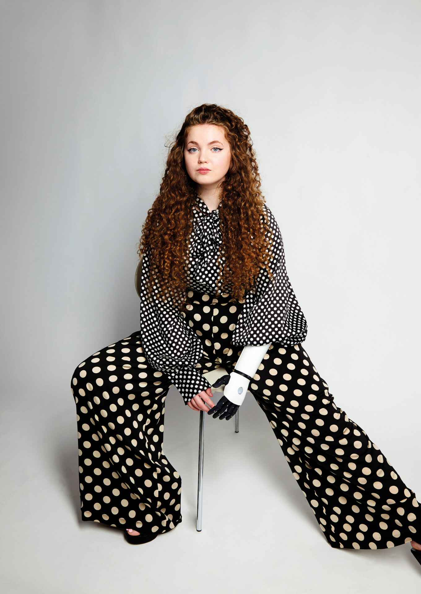 An image of Gracie sitting on a stool. She is wearing a dotted white and black blouse and dotted white and black wide trousers.