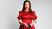 An image of Gracie McGonigal. She has long curly ginger hair and is wearing a red floral dress and red polka dot high heels with a sunflower design. She is using a bionic arm on her left which is decorated with pink sequins.