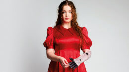 An image of Gracie McGonigal. She has long curly ginger hair and is wearing a red floral dress and red polka dot high heels with a sunflower design. She is using a bionic arm on her left which is decorated with pink sequins.