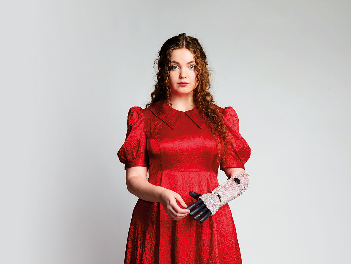 An image of Gracie McGonigal. She has long curly ginger hair and is wearing a red floral dress and red polka dot high heels with a sunflower design. She is using a bionic arm on her left which is decorated with pink sequins.